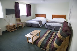 Habitación de hotel con dos camas individuales, un sofá, una mesita de madera, una silla, un televisor montado en la pared y una ventana con cortinas.