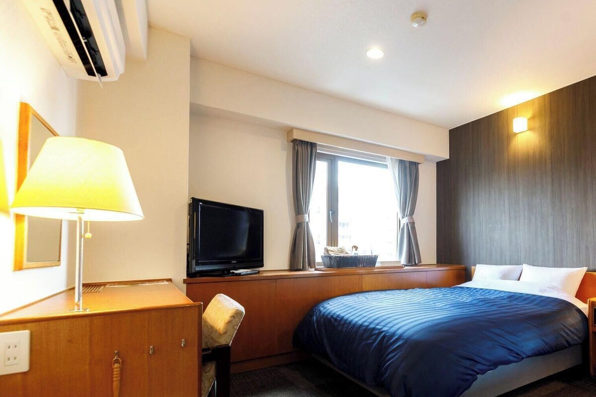 Habitación de hotel con cama, televisión, lámpara, ventana con cortinas y aire acondicionado instalado en la pared.