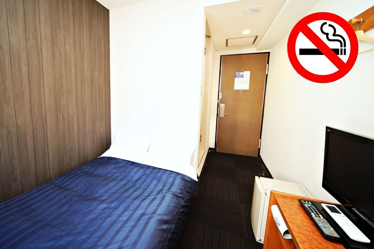 Una habitación de hotel con cama, escritorio, televisión y mando a distancia, un cartel de prohibido fumar y una puerta.