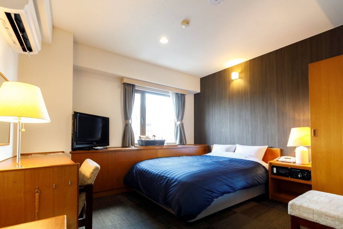 Quarto de hotel com uma cama grande, escrivaninha com luminária, televisão e janela com cortinas.