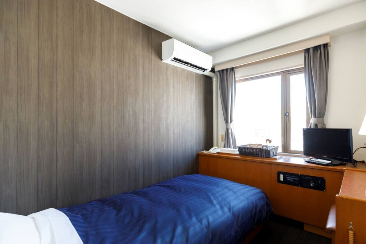 Quarto de hotel com uma cama, uma escrivaninha com computador, ar condicionado e uma janela com cortinas.