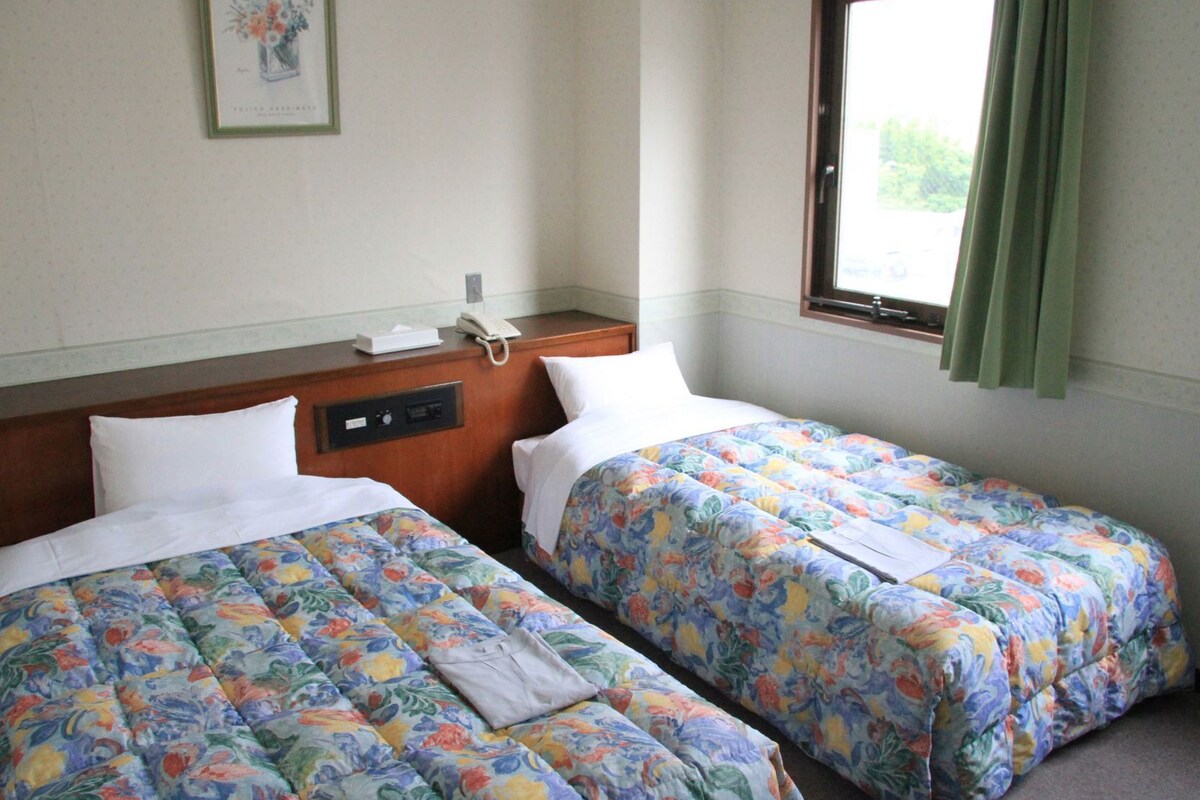 Habitación de hotel con dos camas, una mesita de noche con teléfono, un cuadro en la pared y una ventana con cortinas.