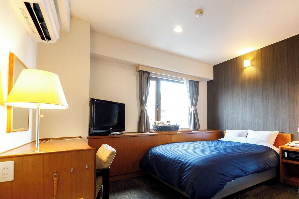 Habitación de hotel con cama, escritorio, televisión, lámpara, ventana con cortinas y luz en la pared.