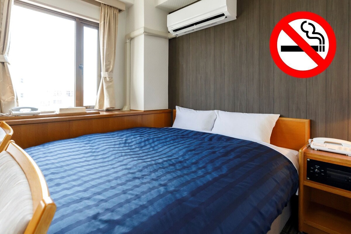 Habitación de hotel con cama, mesita de noche con teléfono, ventana con cortinas y unidad de aire acondicionado.