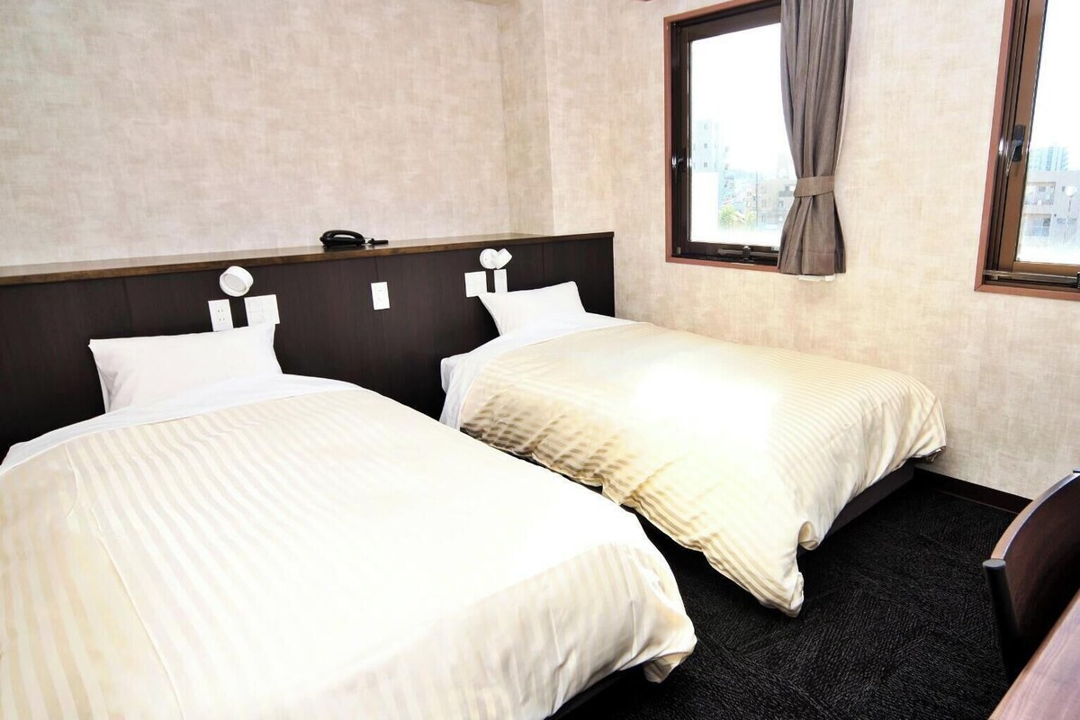 Habitación de hotel con dos camas, una ventana con cortinas, un teléfono y vistas a edificios.