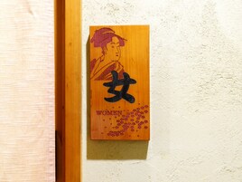Una placa de madera con caracteres japoneses y la palabra 'WOMEN' en una pared con textura.