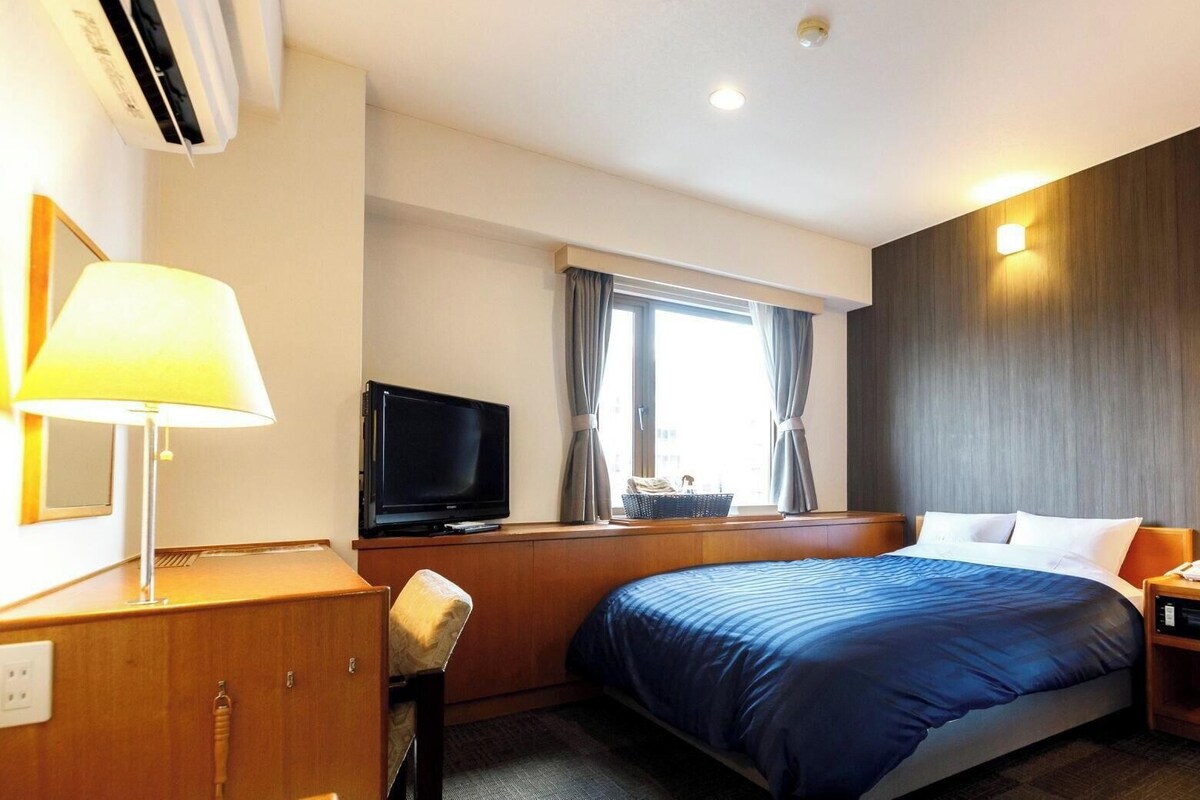 Una habitación de hotel con cama, escritorio con lámpara, televisor y ventana con cortinas.