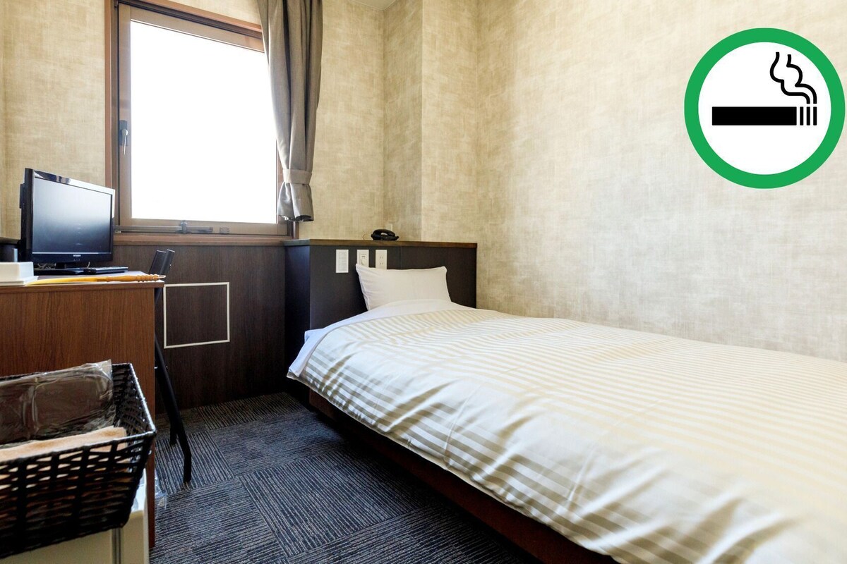 Habitación de hotel con cama, escritorio con computadora, ventana y cartel de prohibido fumar.