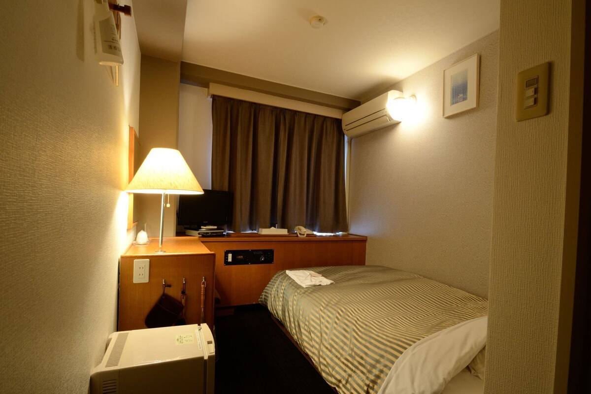 Quarto de hotel com cama, escrivaninha com luminária, televisão, ar condicionado e telefone.
