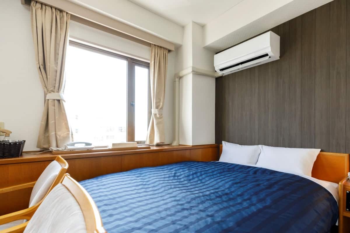 Quarto de hotel com uma cama grande, cabeceira de madeira, janela com cortinas, unidade de ar condicionado e mesa de cabeceira.