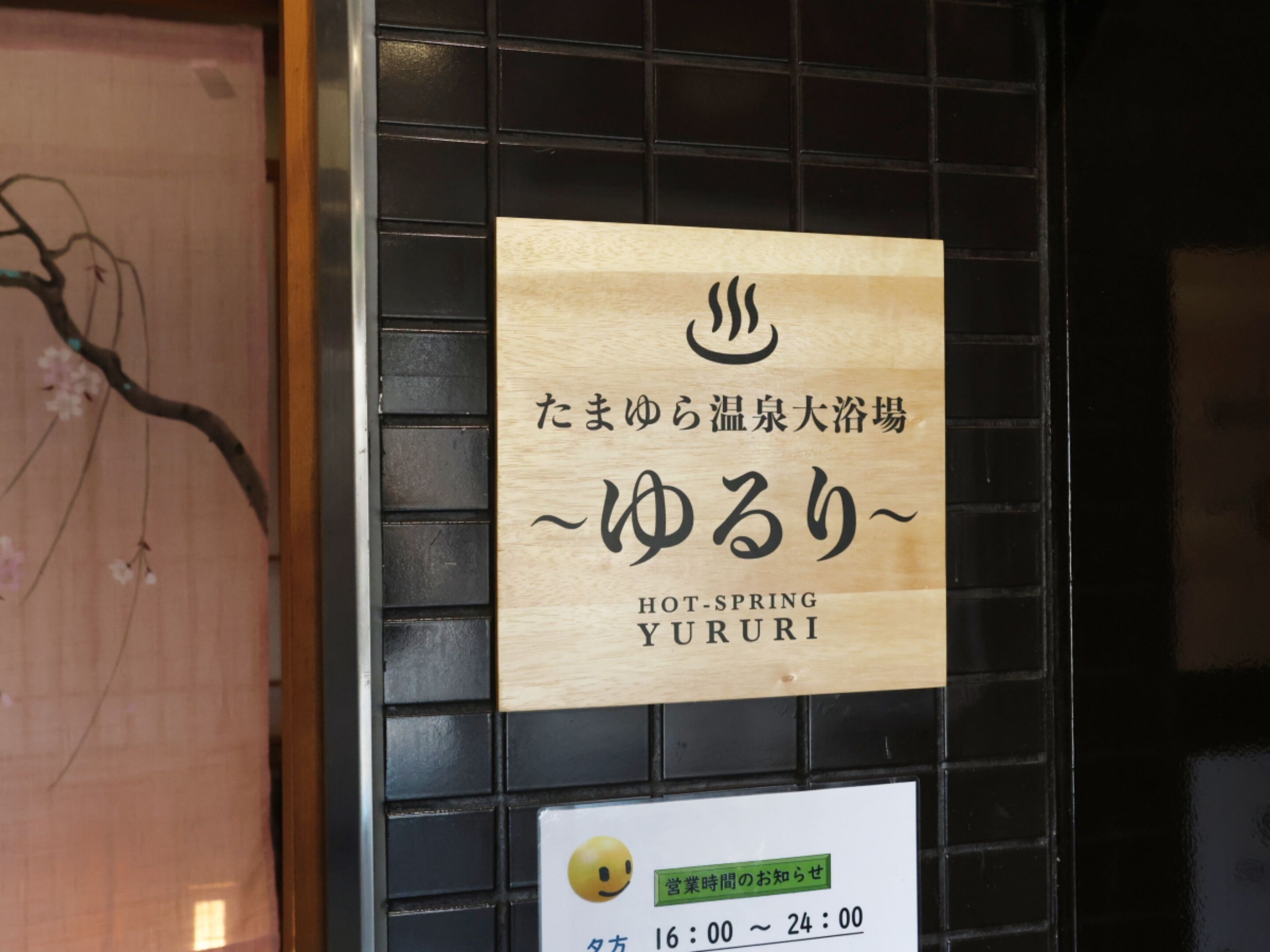 Un letrero con texto japonés y la traducción al inglés 'HOT-SPRING YURURI' en una pared de baldosas negras.