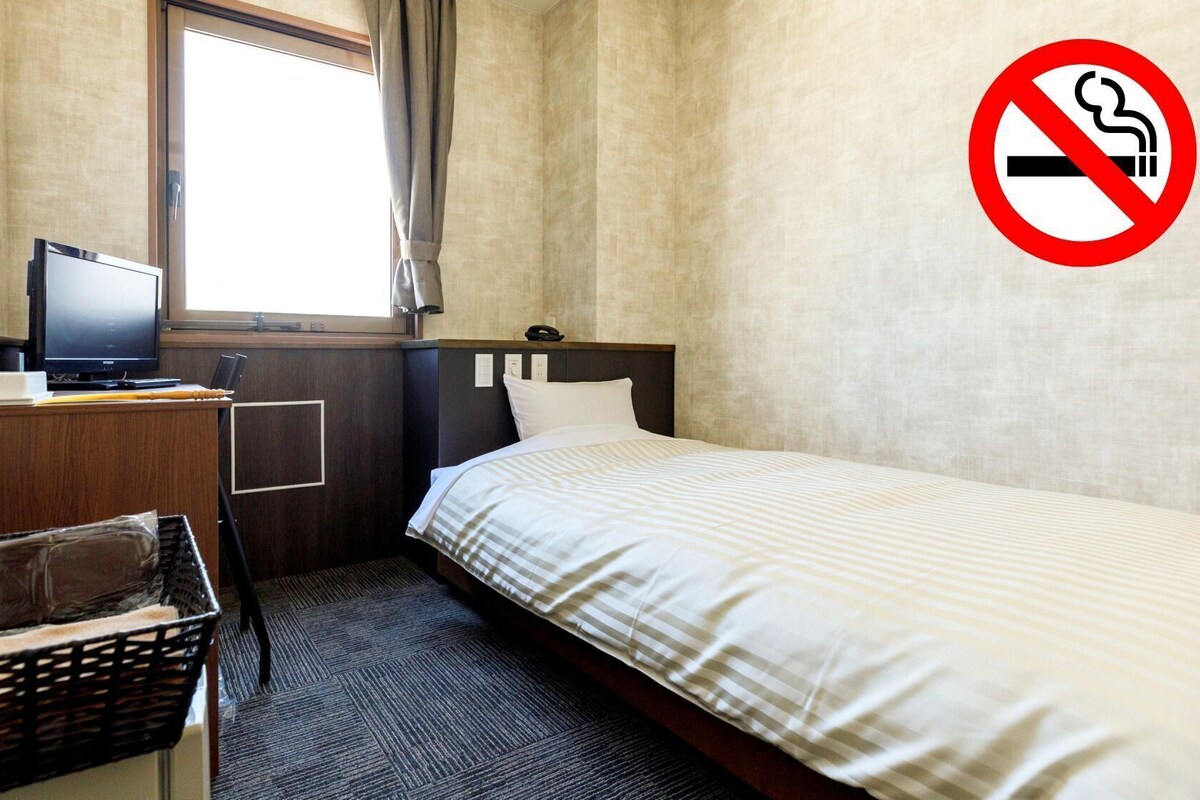 Habitación de hotel con una cama, un escritorio con ordenador, teléfono, ventana y cartel de prohibido fumar.