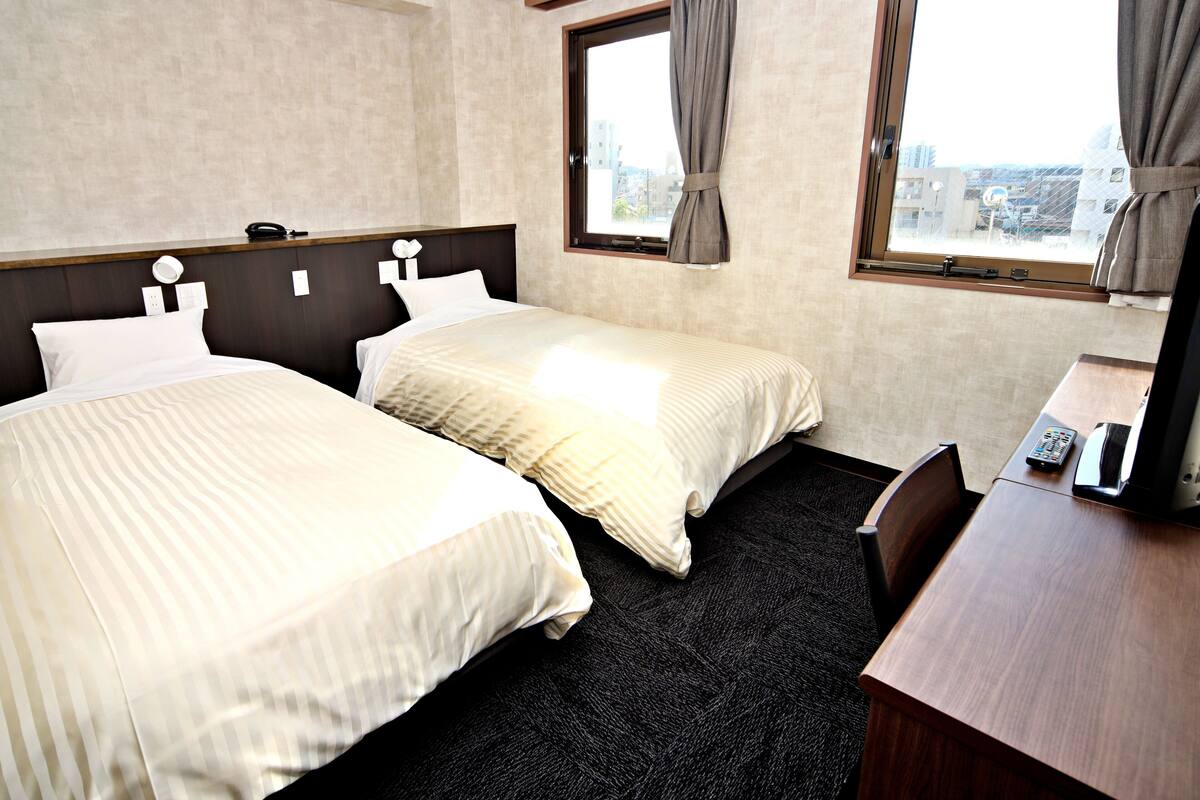 Quarto de hotel com duas camas, uma mesa e uma janela com cortinas.