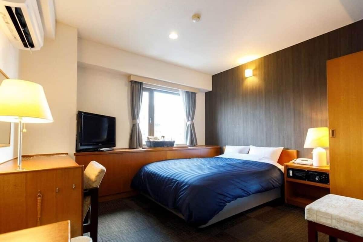 Quarto de hotel com uma cama grande, uma escrivaninha com abajur, televisão e uma janela com cortinas.