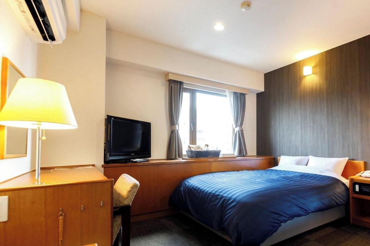 Habitación de hotel con cama, escritorio con lámpara, televisor, ventana con cortinas y luz en la pared.