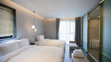 A hotel room with two beds, a bedside table, a lamp, a window with blinds, a bathroom with a glass door, and a view of the city.