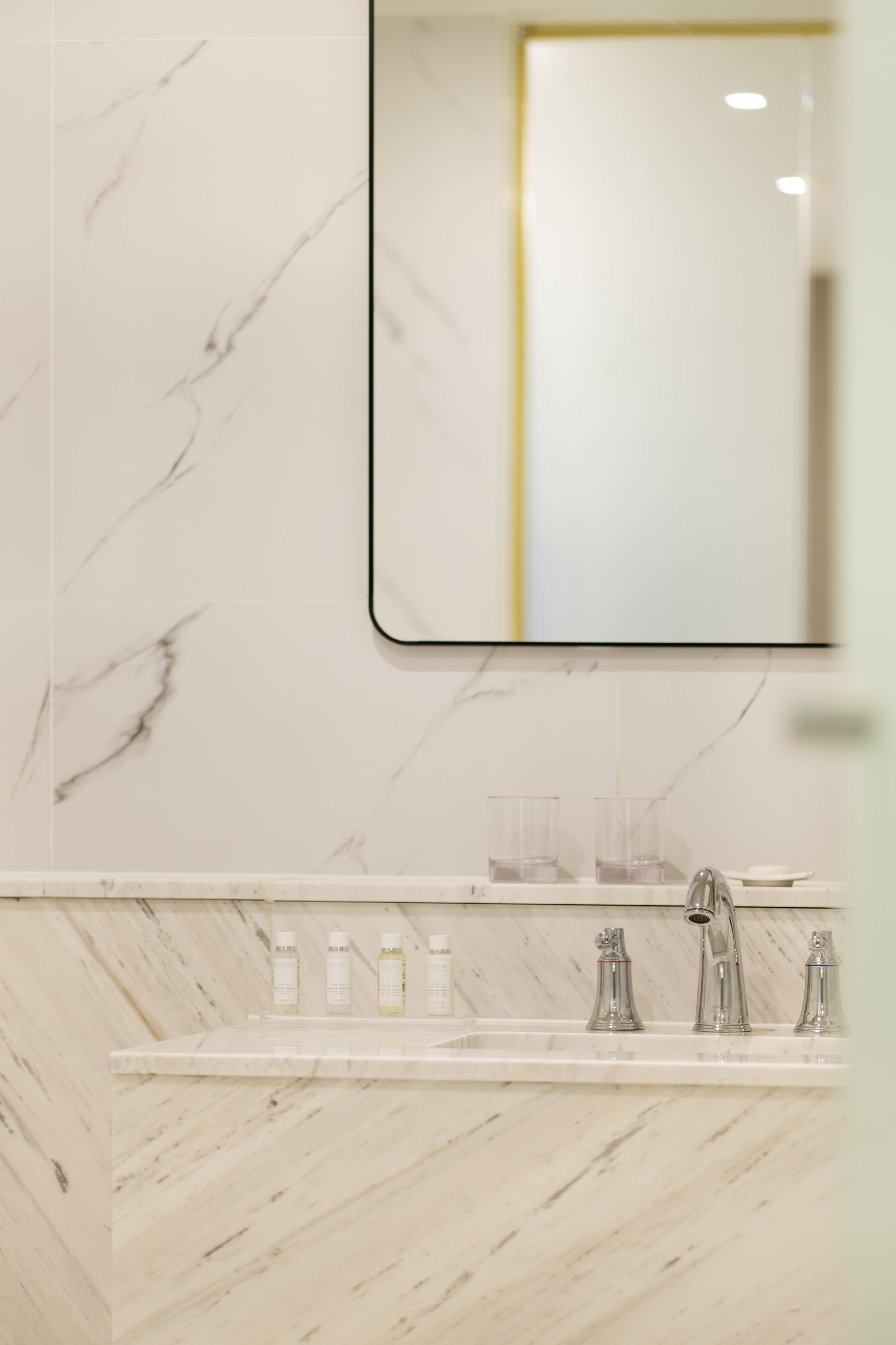 A bathroom with a marble wall, a mirror, and a sink with three faucets and several bottles.