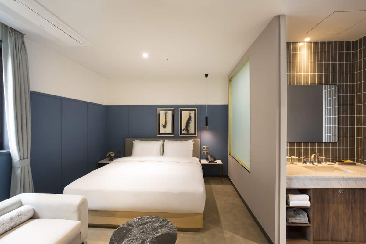 A modern hotel room with a large bed, bedside tables, a desk, and a bathroom with a large mirror and sink.