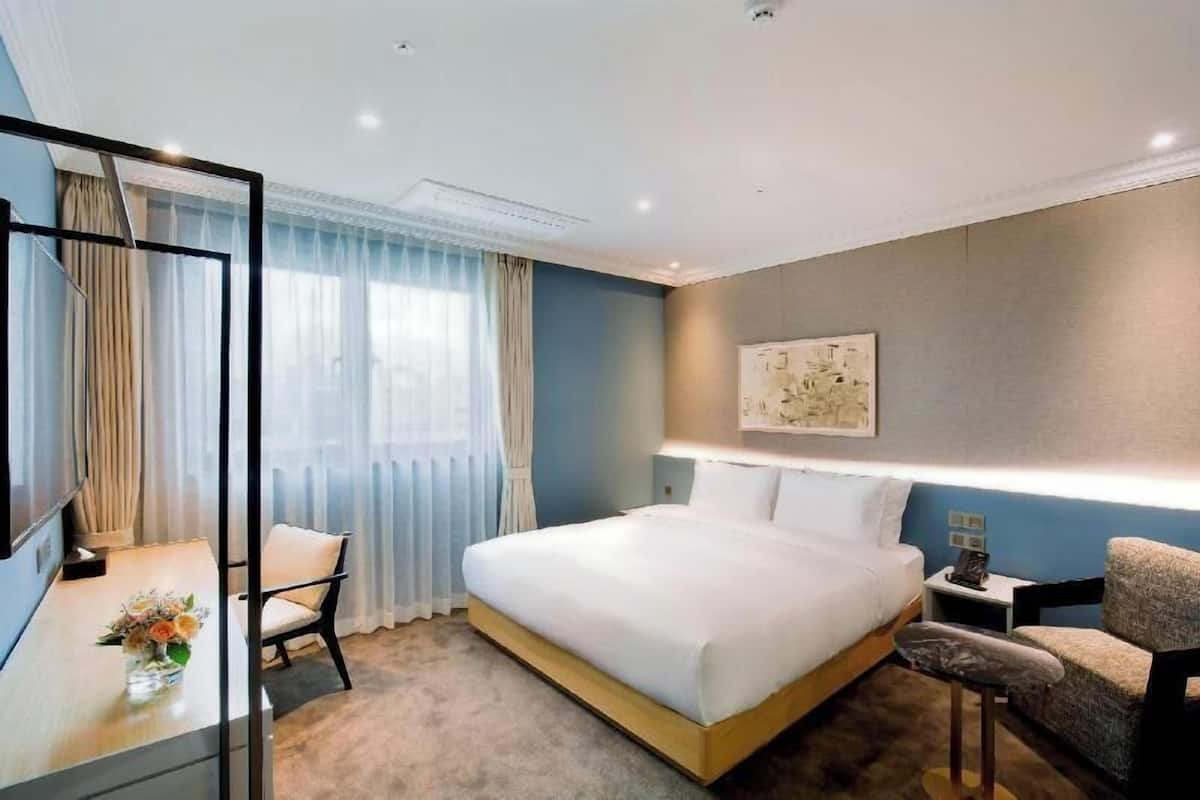 A hotel room with a large bed, a desk with a chair, a small table with a vase, and a wall-mounted TV.