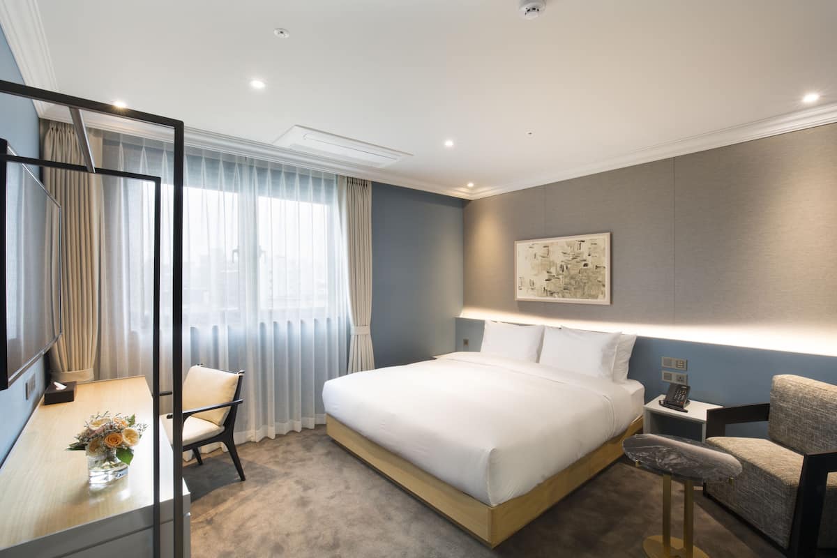 A modern hotel room with a large bed, a desk with a chair, a small table with a vase of flowers, and a large window with curtains.