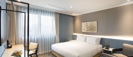 Una habitación de hotel moderna con una cama grande, un escritorio con una silla, una mesita con un jarrón de flores y un ventanal con cortinas.