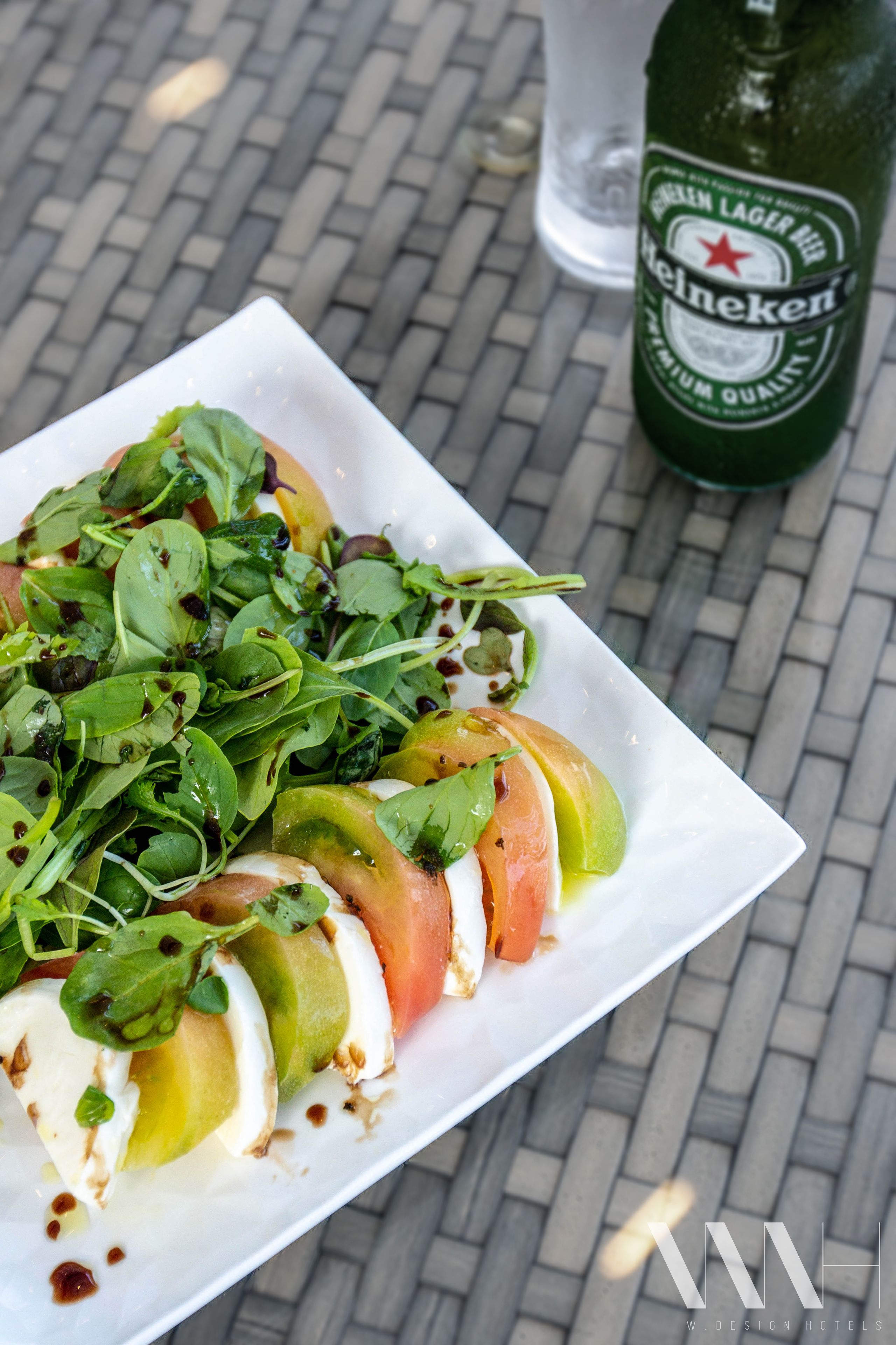A plate of fresh salad with greens and tomatoes, a bottle of Heineken beer, and a glass of water on a brick table.