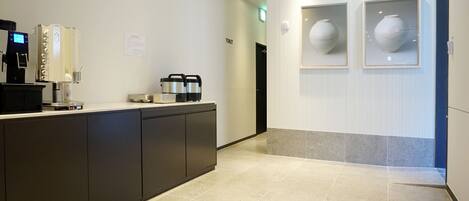 A modern hotel lobby with a reception desk, coffee machines, and framed vases on the wall.