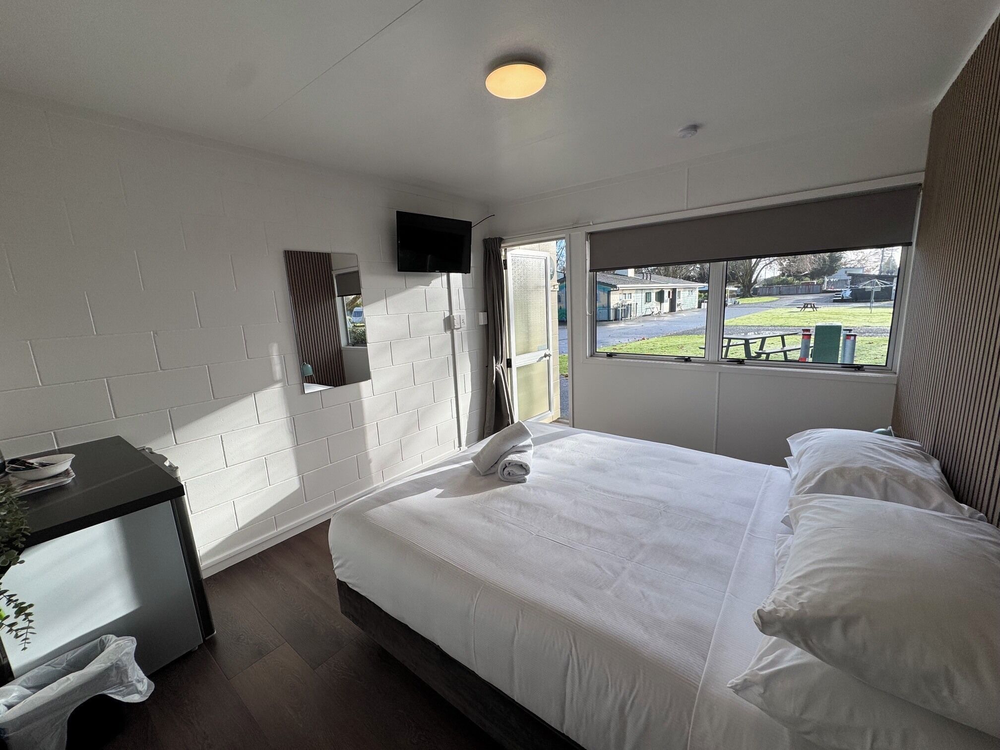 A bedroom with a bed, a bedside table, a mirror, a TV, and a window with a view of the outside.