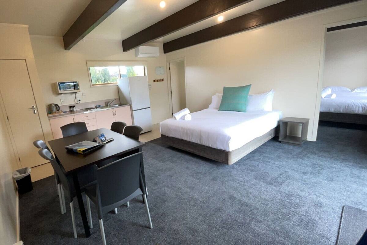 A bedroom with a bed, a desk with chairs, a kitchenette, and an air conditioning unit.