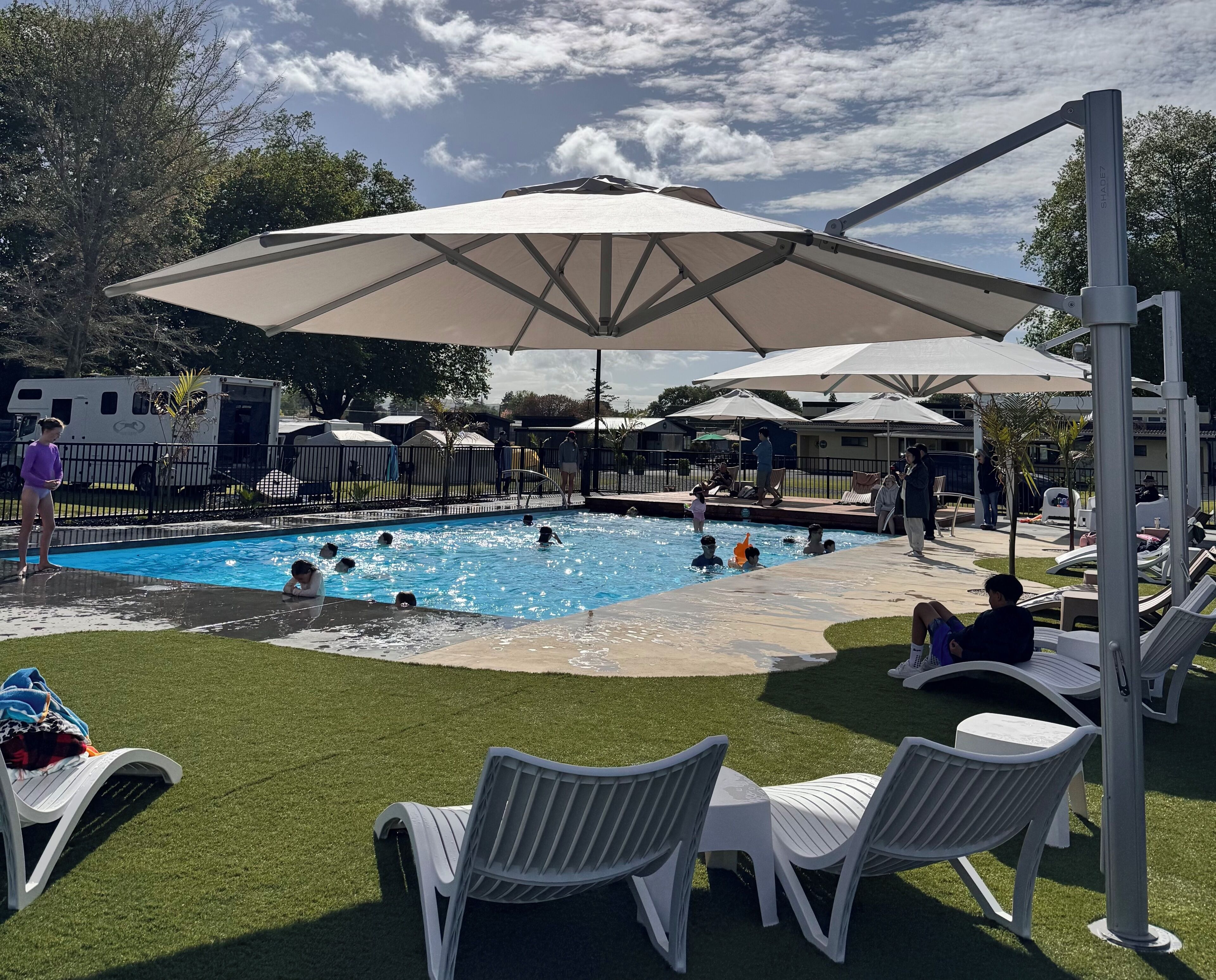 Piscina con sillas de descanso y sombrillas, rodeada por un área vallada con árboles.