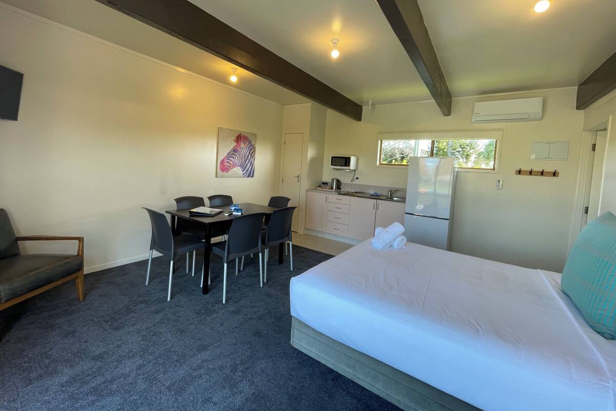 A room with a bed, a dining table with chairs, a kitchen area, and a ceiling fan.