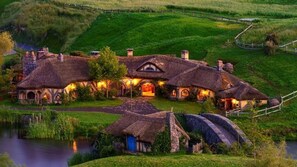 A Hobbiton-style house with multiple thatched-roof structures, a central building with a large door, and a surrounding landscape with a pond and grassy hills. - Cambridge TOP 10 Holiday Park (Cambridge)