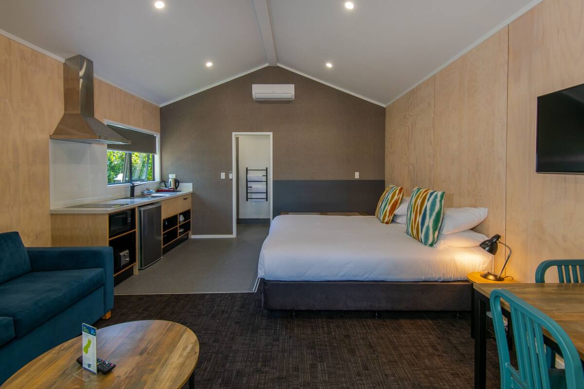 A modern hotel room with a kitchenette, a large bed with multiple pillows, a sofa, and a dining area.