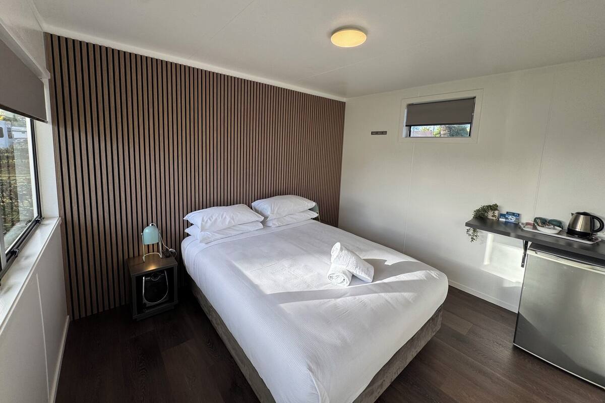 A hotel room with a bed, bedside table, lamp, and a kitchenette area.