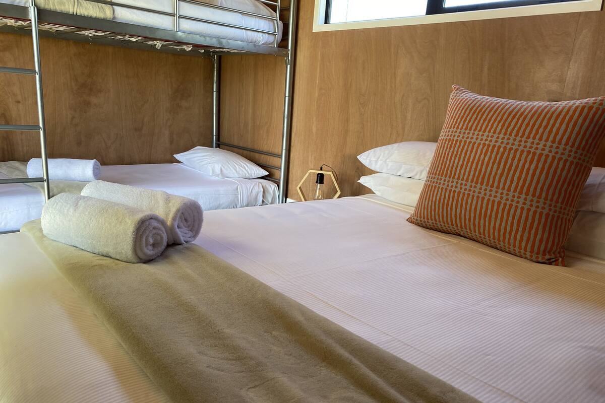 A hotel room with a bunk bed, a single bed with a beige blanket, white pillows, and a patterned orange pillow.