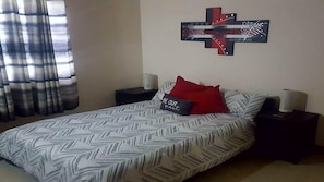 A bedroom with a bed, a cross-shaped wall decoration, a pillow with text, and two bedside tables with lamps.