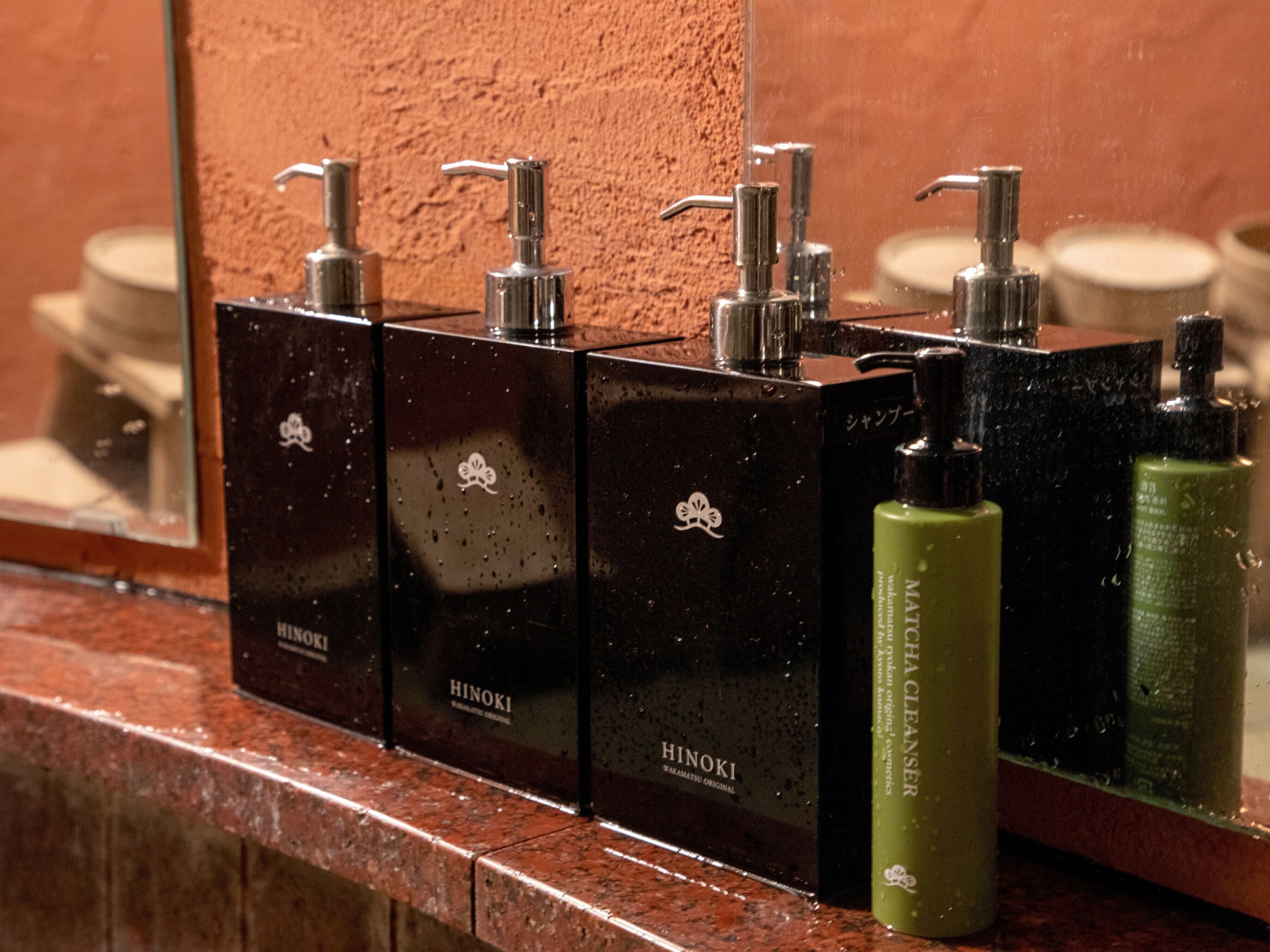A bathroom with a mirror reflecting a row of black toiletries dispensers labeled "Hinoki".