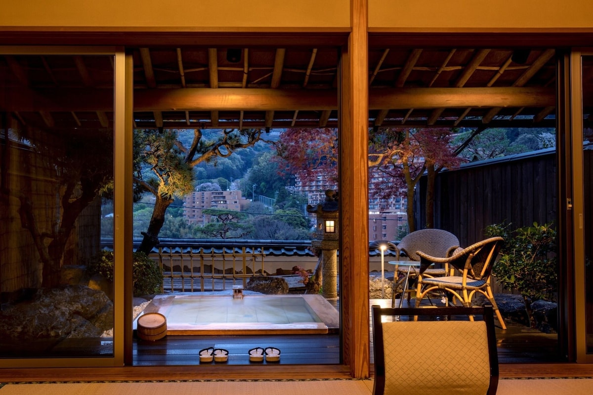 Ett traditionellt japanskt rum med tatamimattor på golvet, ett träpanelat tak och utsikt över en trädgård med en damm och träd.