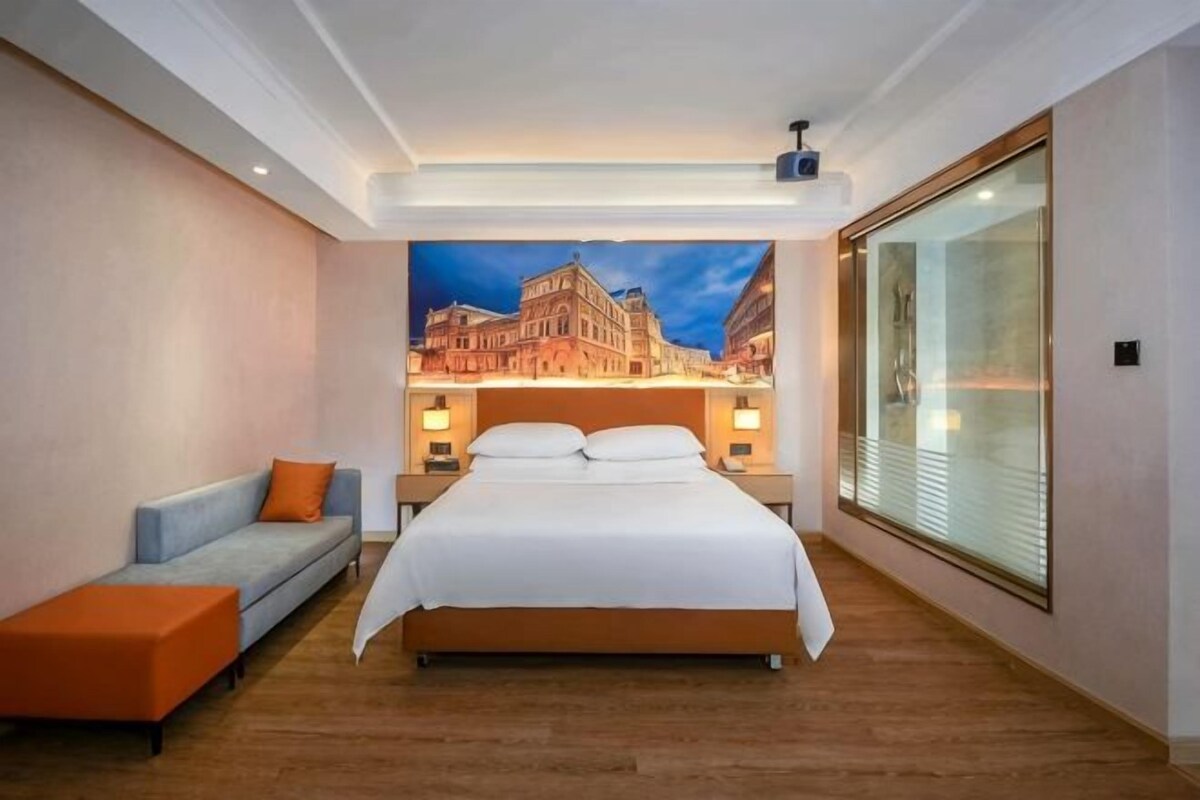 A hotel room with a large bed, a sofa, a nightstand with lamps, and a cityscape mural on the wall.