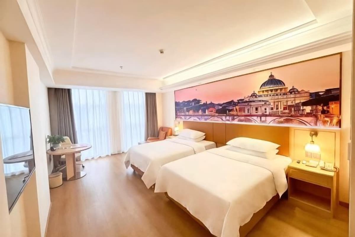 A hotel room with two beds, a desk, a chair, a TV, and a painting of a cityscape.