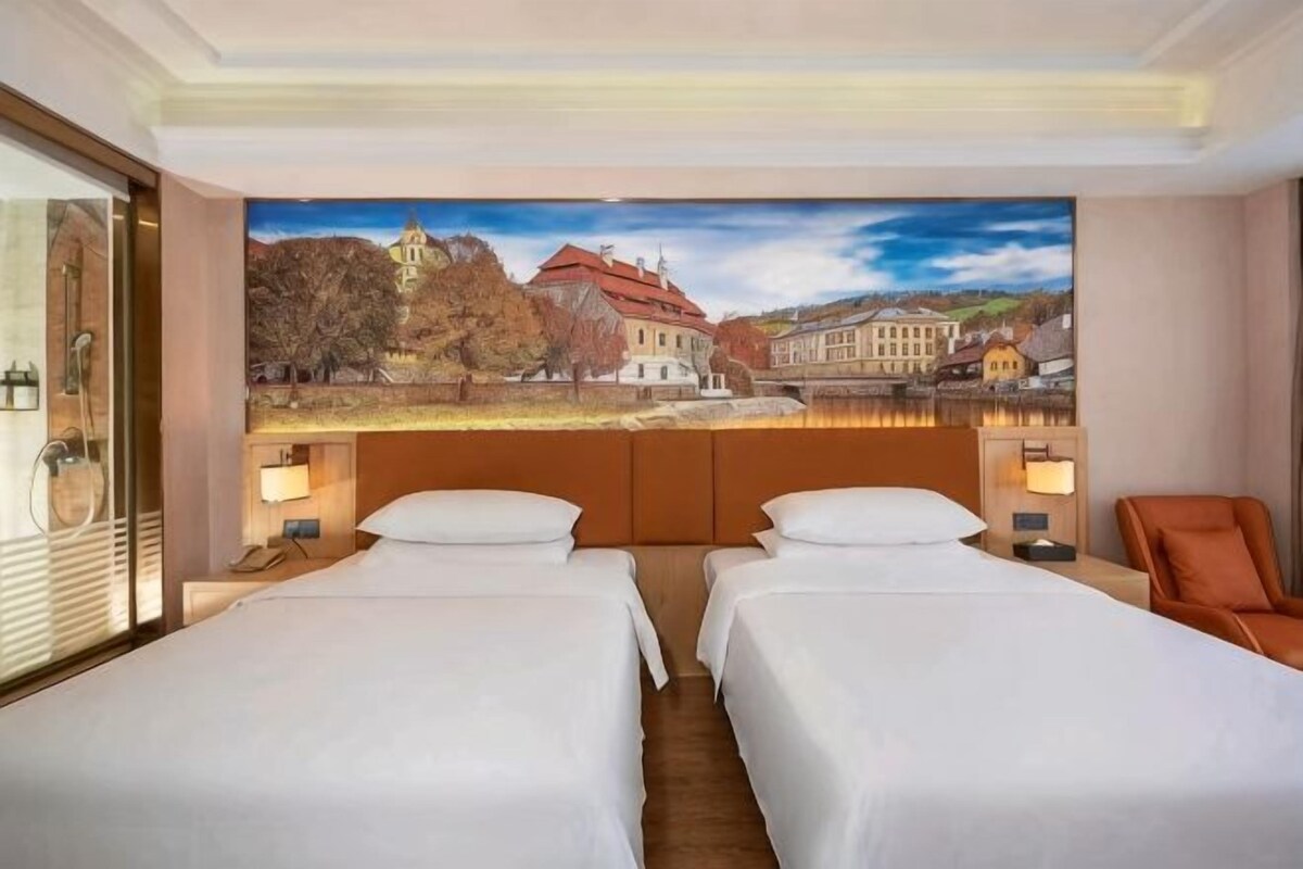 A hotel room with two beds, a large window with a scenic view, and a painting above the beds.