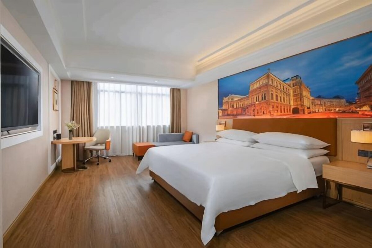 A hotel room with a large bed, a desk, a chair, and a painting of a building.