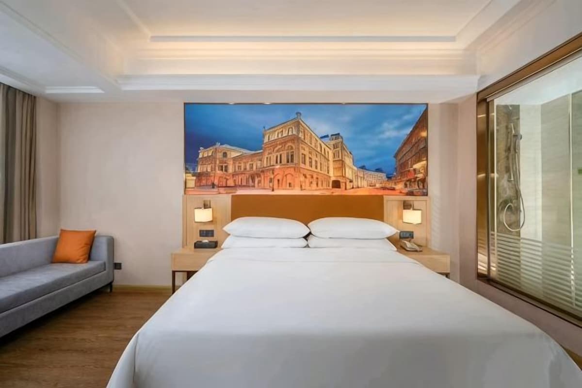 A hotel room with a large bed, a sofa, a nightstand with a lamp, and a painting of a building on the wall.