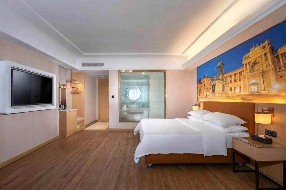 A hotel room with a bed, bedside table, TV, and a painting of a building.