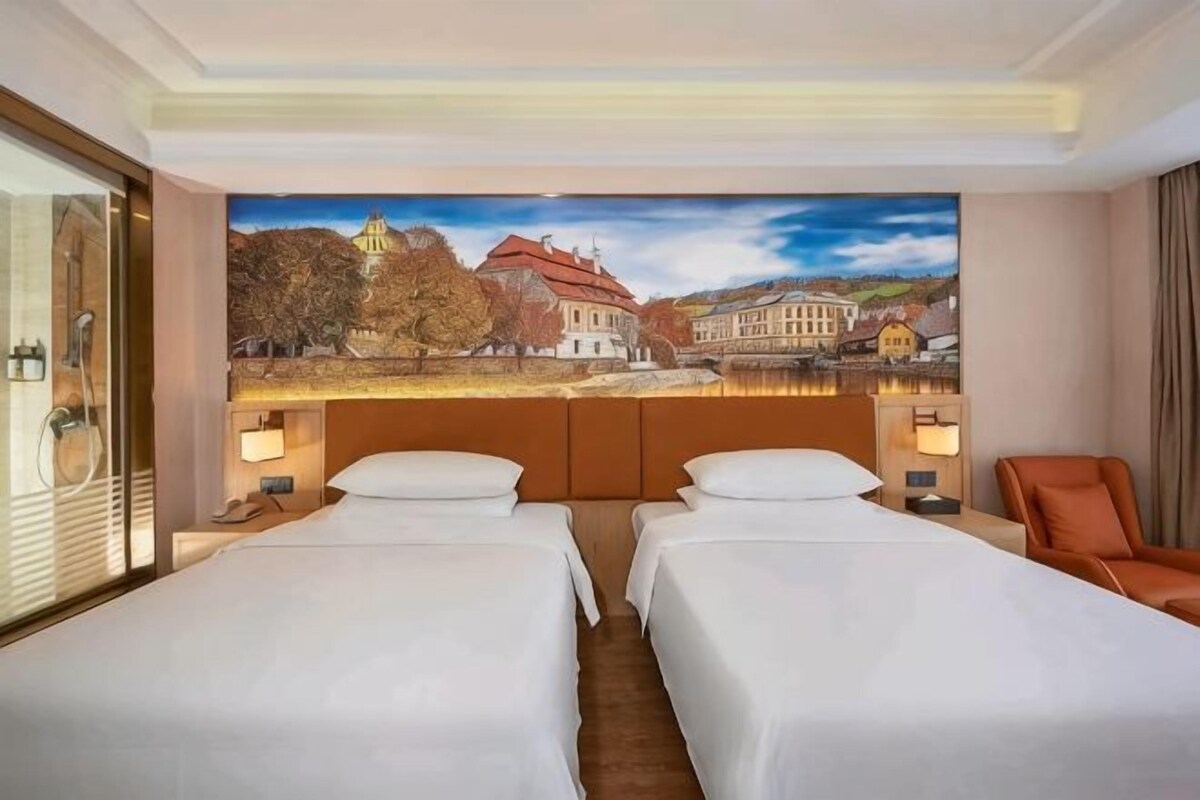A hotel room with two beds, a large window, and a view of a cityscape.
