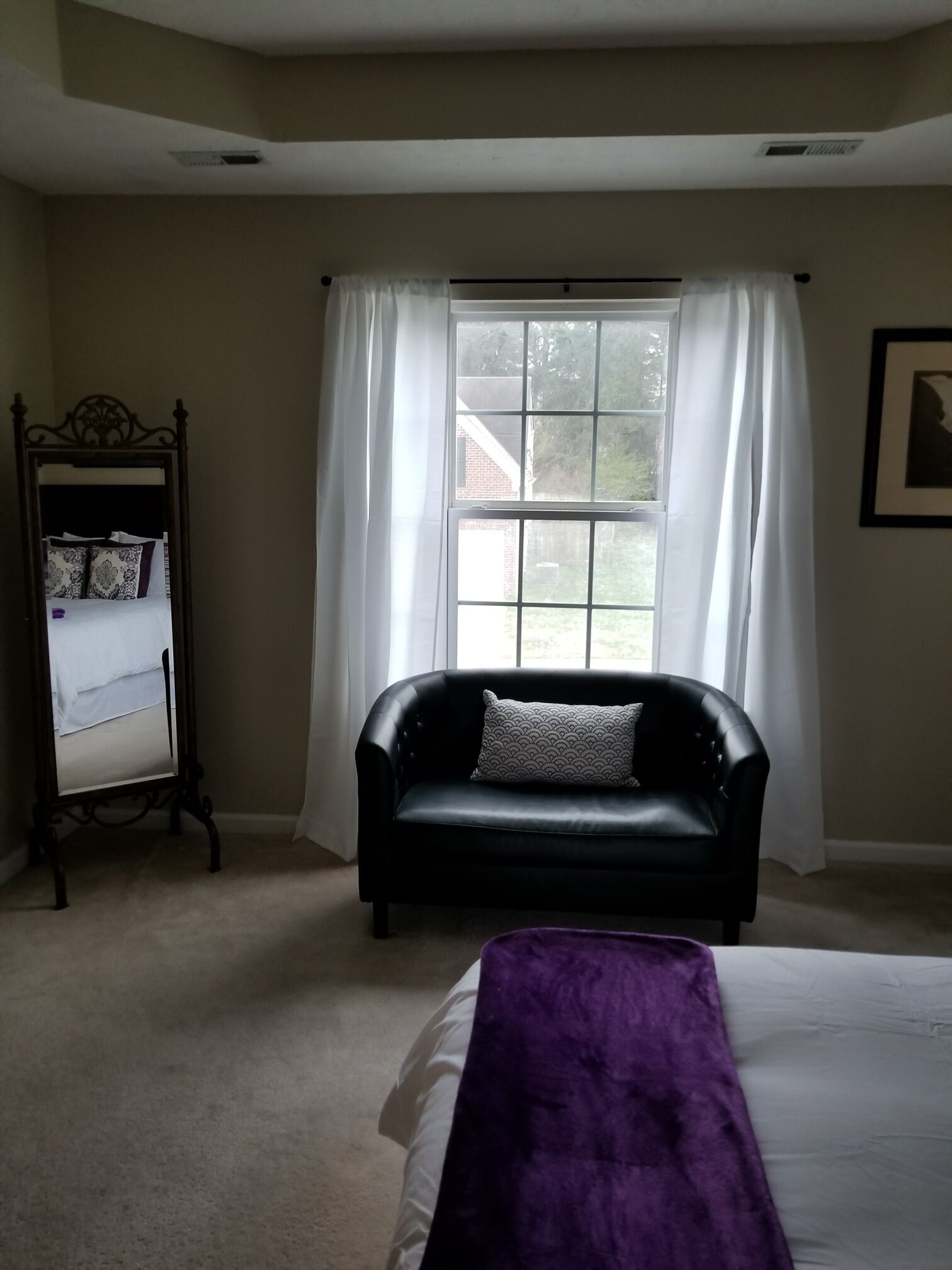 A bedroom with a black leather sofa, a bed with a purple blanket, a standing shelf with decorative pillows, and a large window with white curtains.