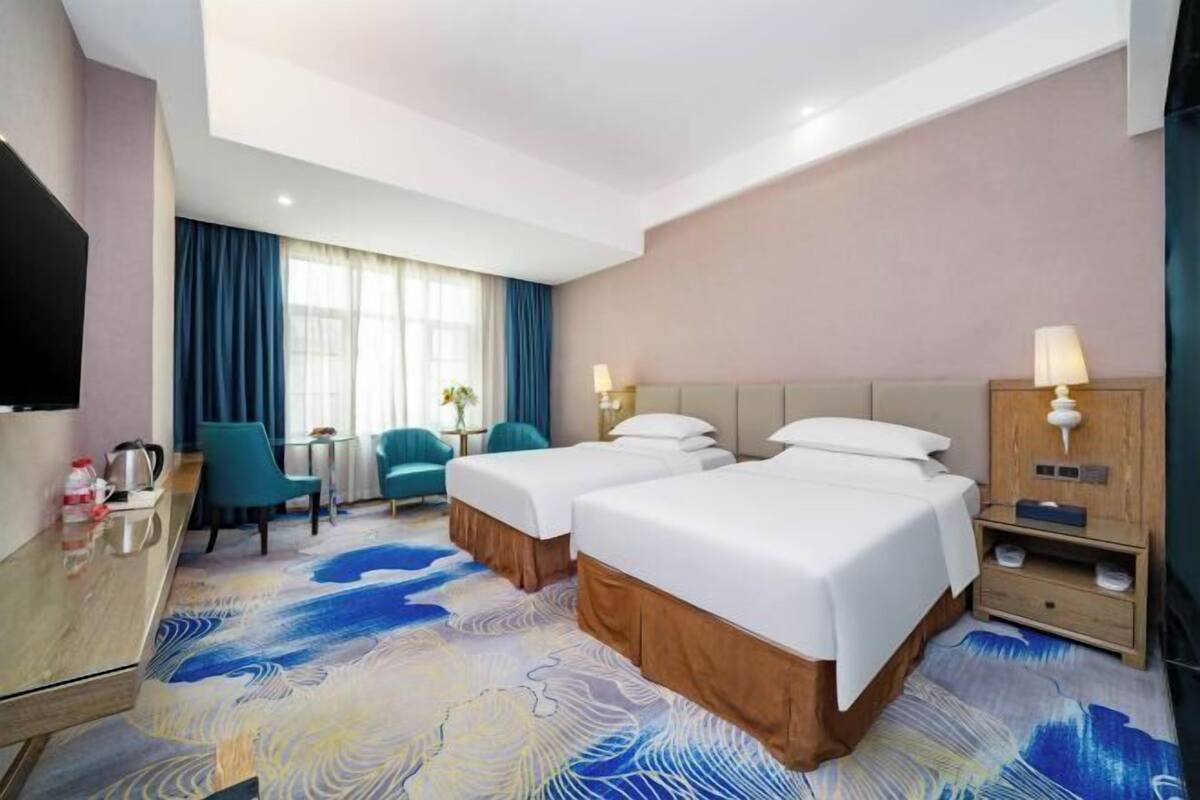 A hotel room with two beds, a desk, a chair, a TV, and a lamp.