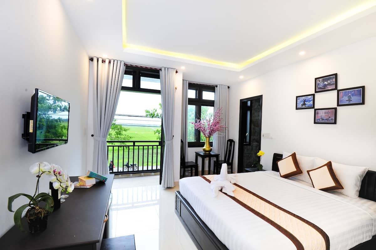 A hotel room with a bed, a desk with a TV, a balcony with curtains, and framed pictures on the wall.
