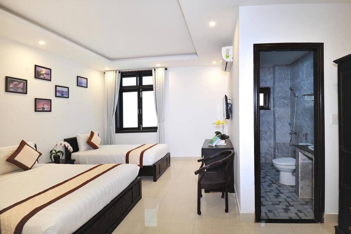 A hotel room with two beds, a desk, a chair, a window with curtains, and a bathroom visible through an open door.