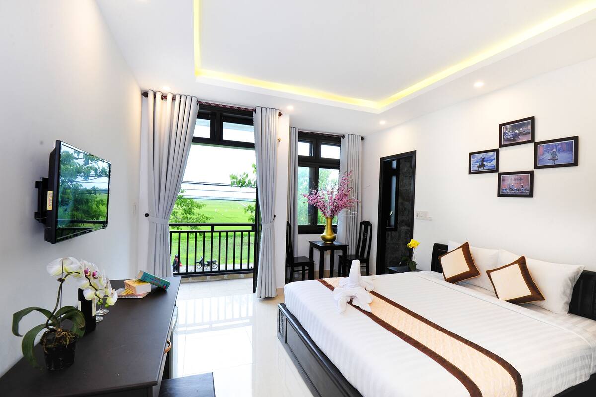 A hotel room with a bed, a desk, a television, and a balcony with a view of greenery.